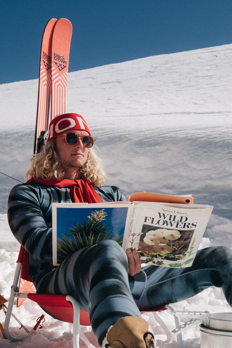 Man sits in Merino thermals on a deck chair reading a book in the snow on a bluebird day