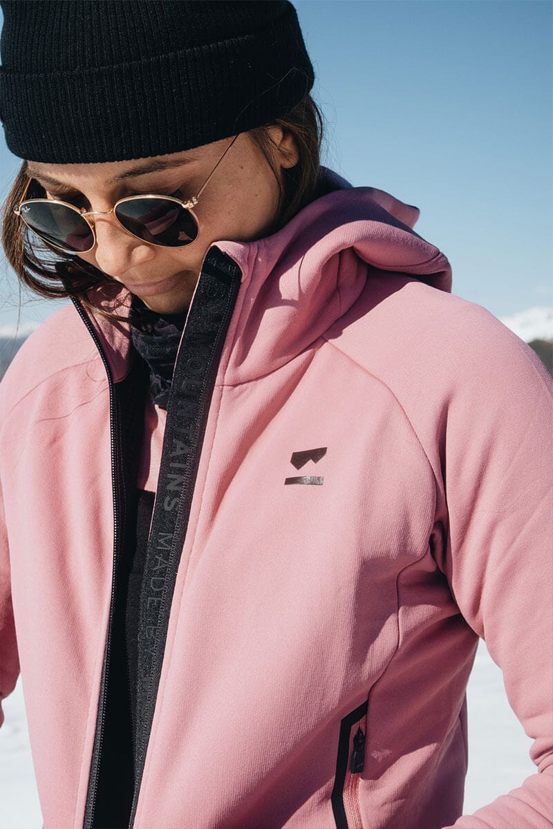 Women in the snow wearing beanie and glasses and zipping up a pink Merino mid-layer