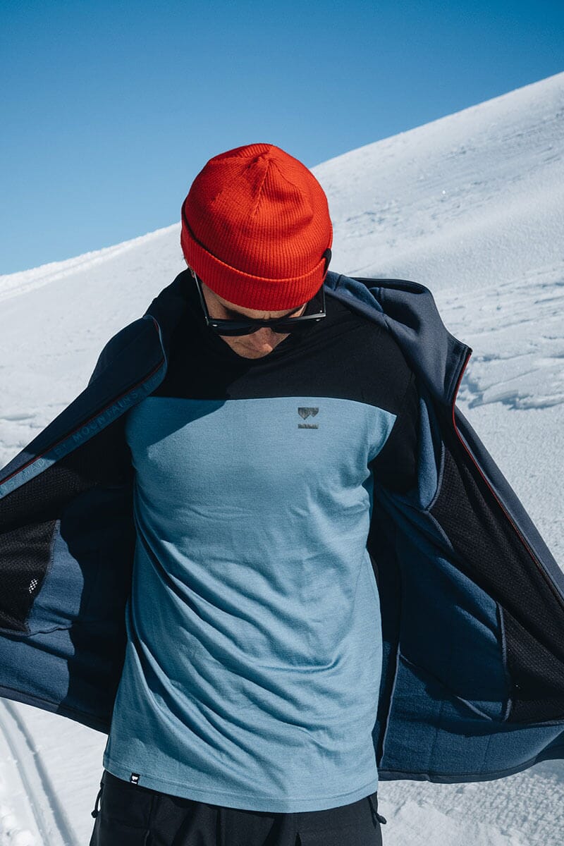 Man standing in the snow with a beanie on, wearing merino layers and taking off a jacket