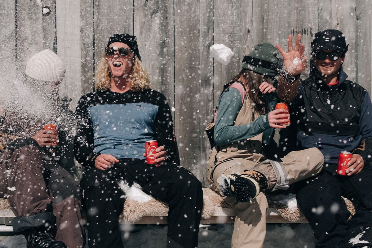 A group of four sit on a bench outside a hut drinking beer and dodging snowballs that are being thrown at them.